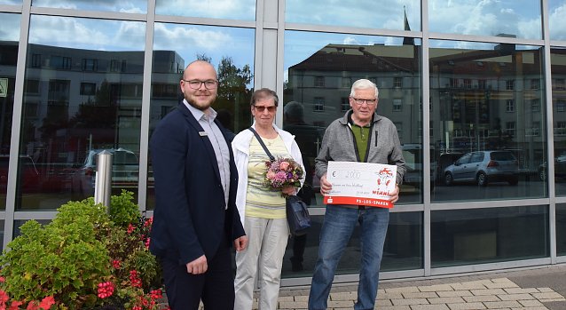 Bild: Kundenberater Jonas Echtermeier &uuml;berreicht den gl&uuml;cklichen Gewinnern einen symbolischen Scheck und Blumen.