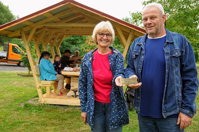 Bild: Ortschaftsb&uuml;rgermeisterin Petra Gerlach und Zimmermann Mario Eckert vor der neuen Waldschenke. Das Schild wurde an der Sitzgruppe angebracht und erinnert daran, dass der Ilfelder Ortschaftsrat diese gesponsert hat. 